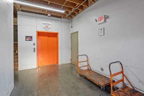 Indoor loading area with orange elevator doors, moving carts, exit sign, and a concrete floor