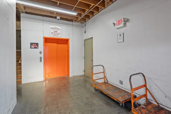 Indoor loading area with orange elevator doors, moving carts, exit sign, and a concrete floor