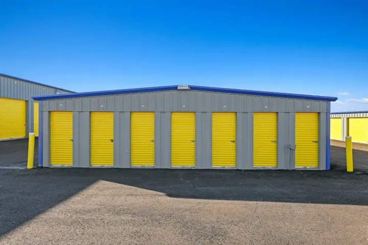 Exterior of a drive-up storage building with gray siding and seven yellow roll-up doors under a blue sky