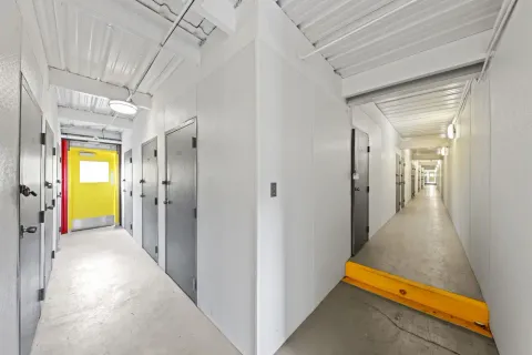 Hallway of indoor storage units in Irving on N O'Connor Rd with gray unit doors, white walls, a yellow exit door, and a ramp