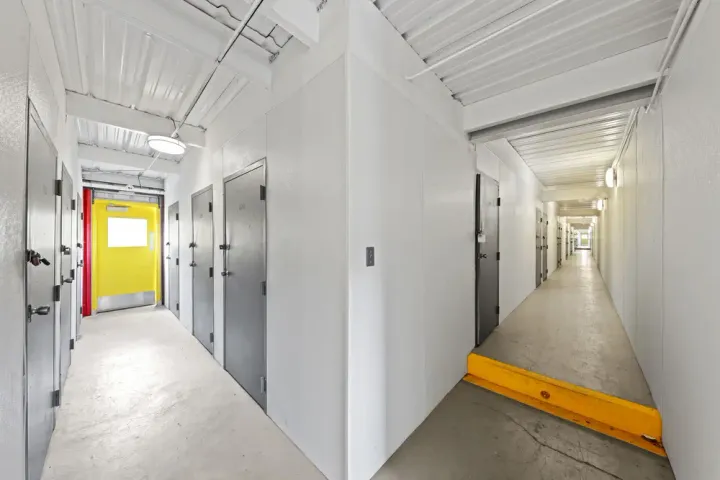 Hallway of indoor storage units in Irving on N O'Connor Rd with gray unit doors, white walls, a yellow exit door, and a ramp