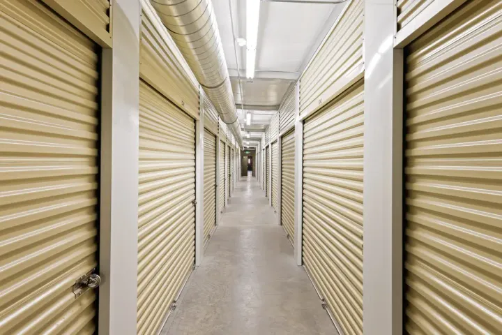 Well-lit interior self-storage hallway with beige roll-up unit doors and overhead ventilation ductwork
