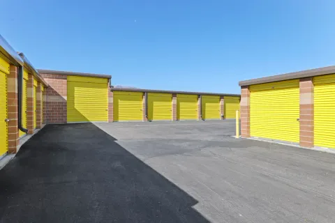 Outdoor drive-up self-storage units with yellow roll-up doors and brick pillars under a clear blue sky