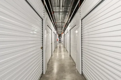 Indoor hallway with white corrugated metal roll-up storage unit doors and exposed ceiling pipes