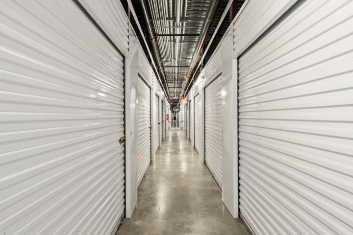 Indoor hallway with white corrugated metal roll-up storage unit doors and exposed ceiling pipes