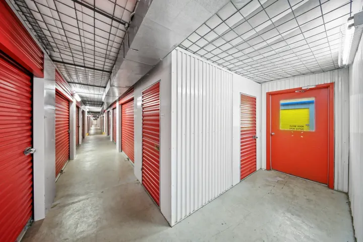 Indoor storage unit hallway with red roll-up doors, white corrugated walls, and wire mesh ceilings